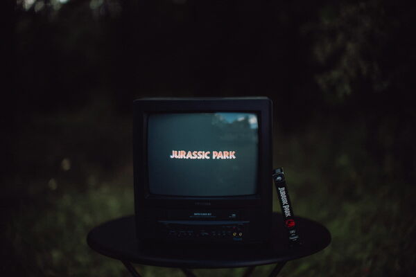 Ein alter Röhrenfernseher auf einem Tisch im Freien, auf dessen Bildschirm der Filmtitel „Jurassic Park“ leuchtet. Daneben steht die passende VHS-Kassette.