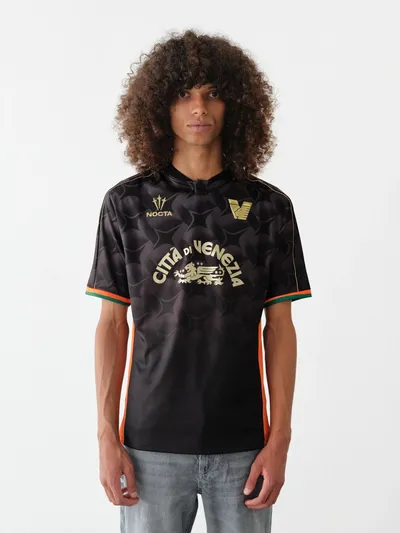 Junger Mann mit lockigem Haar trägt schwarzes FC Venedig Trikot mit goldener Aufschrift 'Città di Venezia' und Logos, dazu Jeans