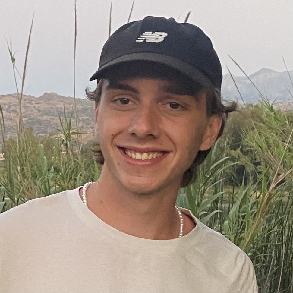 Junger Mann mit schwarzer Kappe, weißem T-Shirt und Perlenkette lächelt vor einer Landschaft mit grünen Pflanzen und Bergen