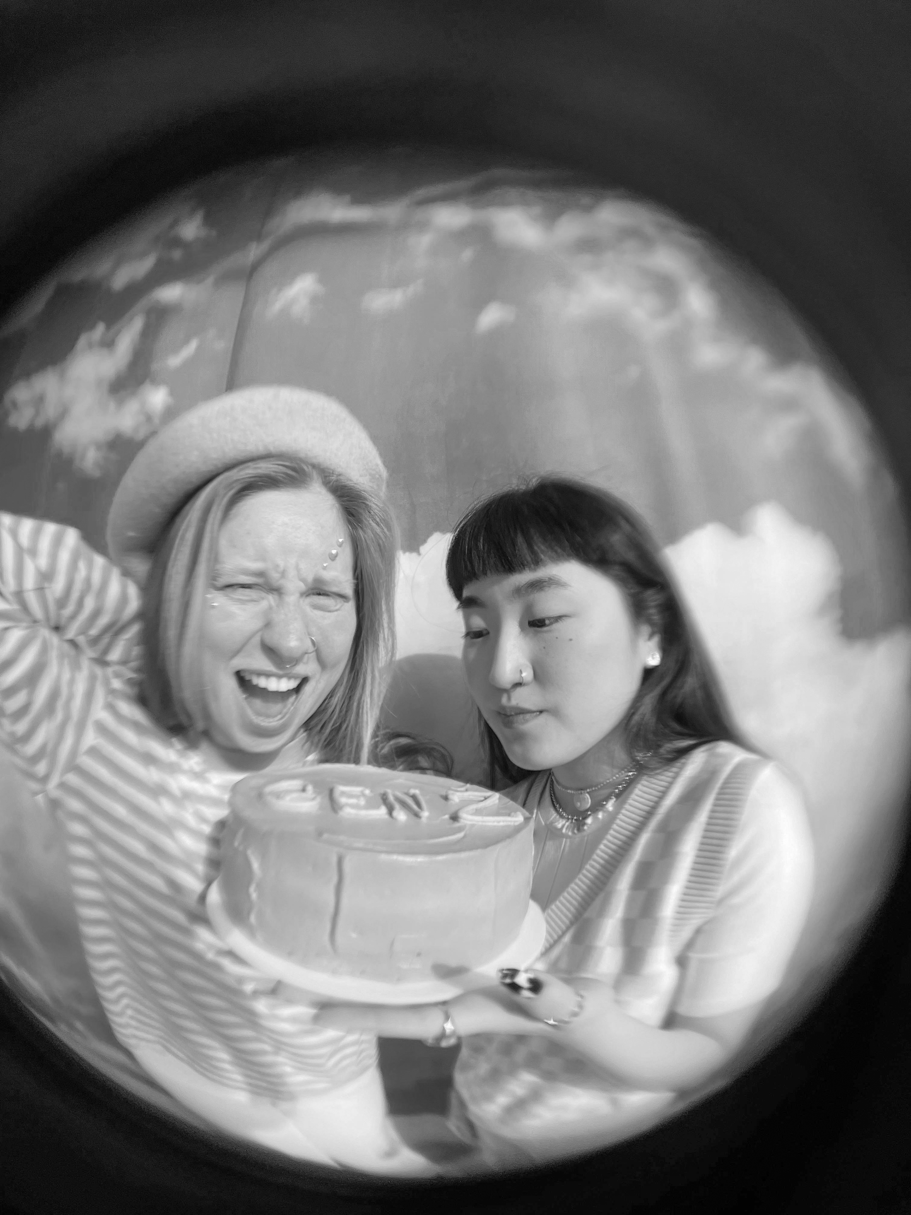 Zwei Frauen halten gemeinsam einen runden Kuchen mit Schriftzug, eine lacht mit geschlossenen Augen, die andere schaut konzentriert auf den Kuchen