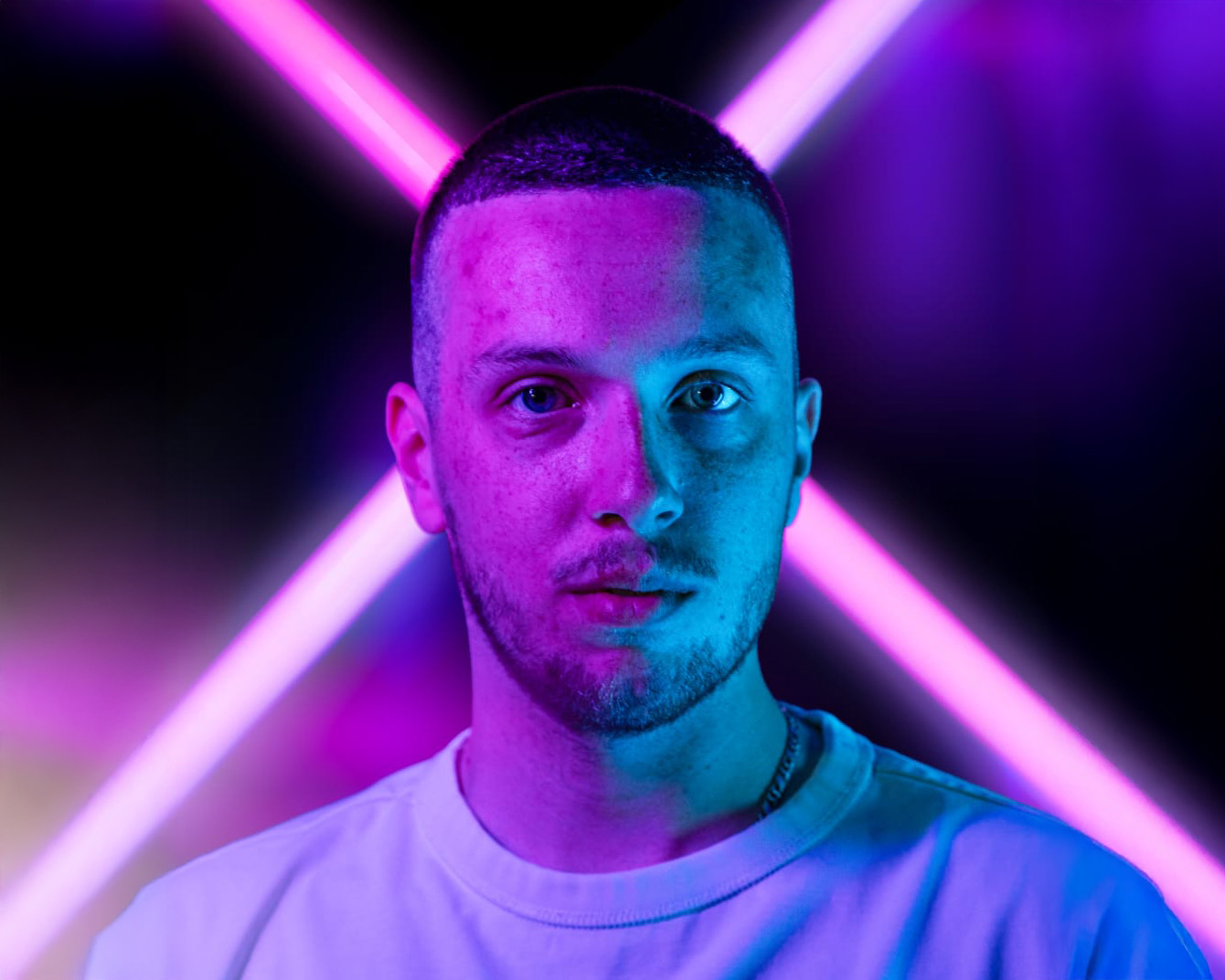 Porträt eines jungen Mannes mit kurzem Haar und Bart, beleuchtet von pink-blauen Neonlichtern vor schwarzem Hintergrund