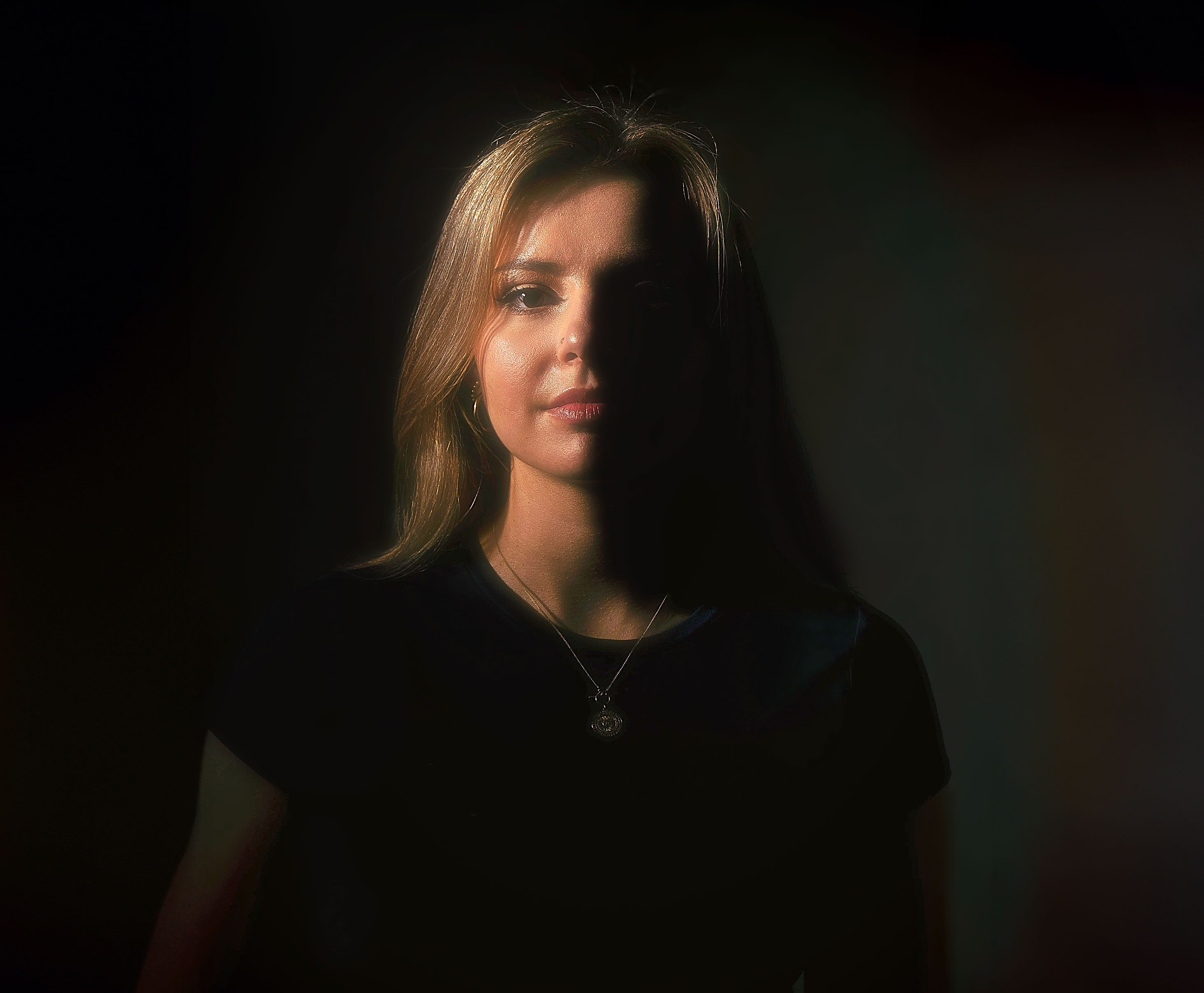 Frau mit langen blonden Haaren, die Hälfte ihres Gesichts ist von Schatten bedeckt, sie trägt ein schwarzes Oberteil und eine Halskette mit Anhänger