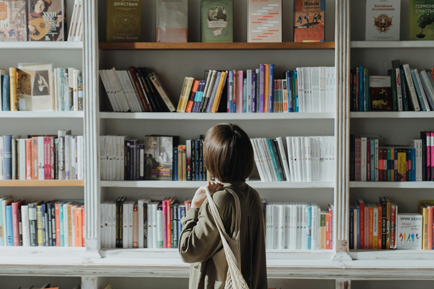Person mit kurzem Haar und grüner Jacke steht vor einem Bücherregal mit bunten Büchern und blickt darauf