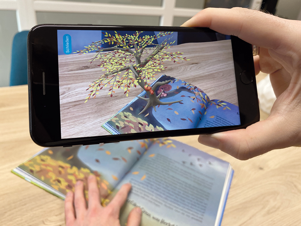 Hand hält Smartphone, das einen animierten Baum mit fallenden Blättern über einem aufgeschlagenen Buch zeigt
