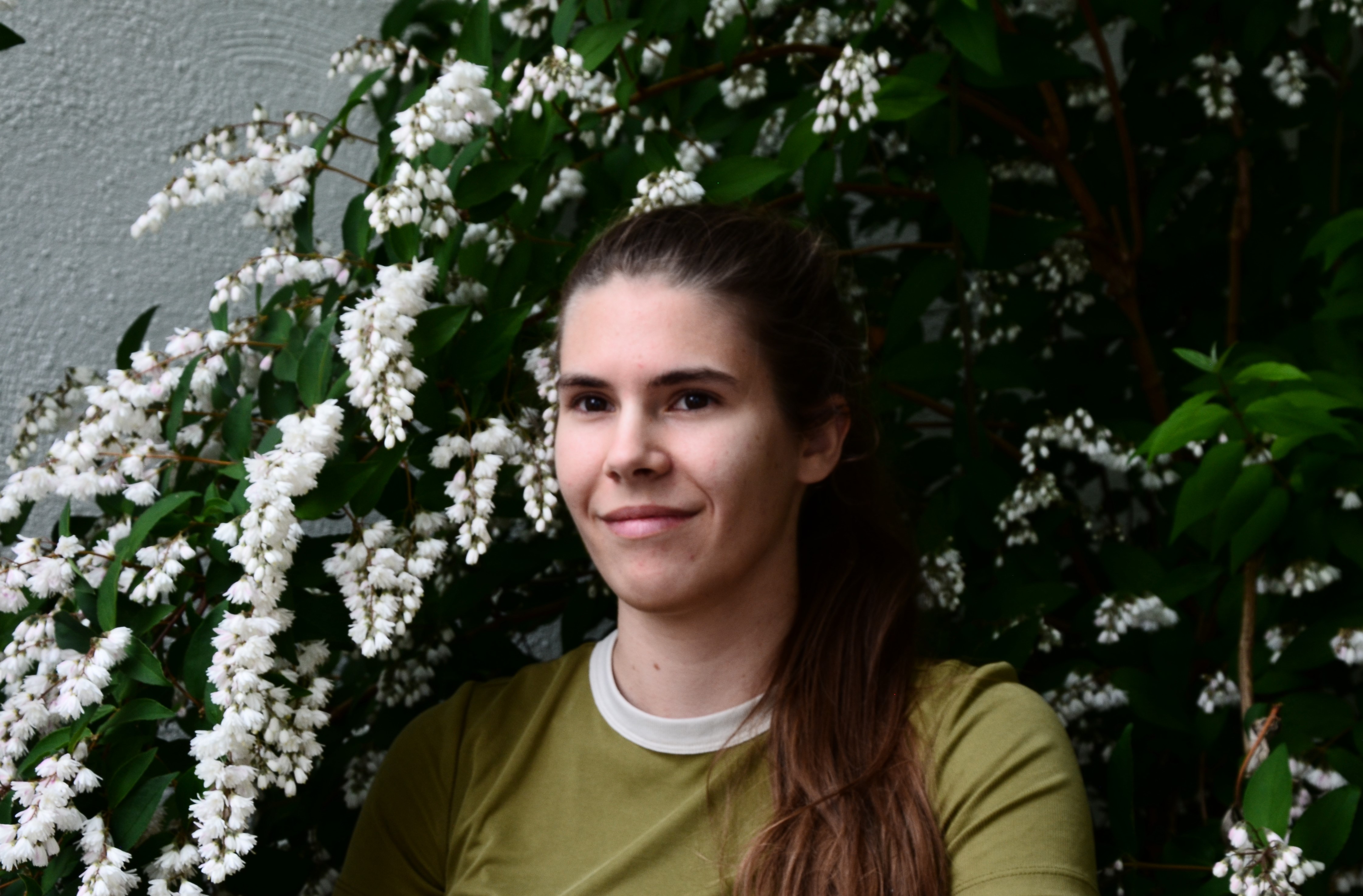 Junge Frau mit langem braunem Haar und grünem Shirt vor einem Strauch mit weißen Blüten