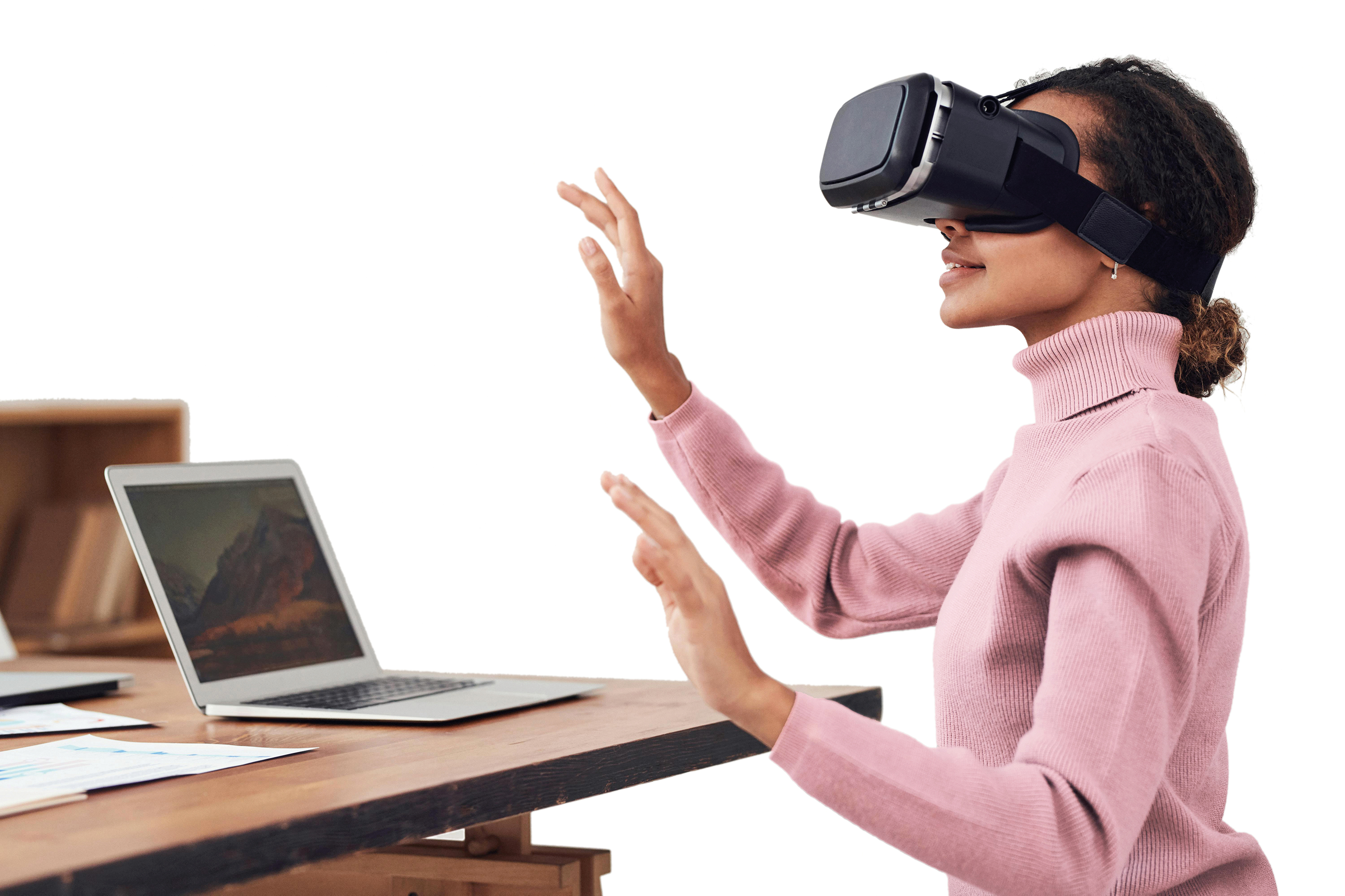 Frau mit rosa Rollkragenpullover nutzt VR-Brille und gestikuliert vor einem Laptop auf einem Holztisch
